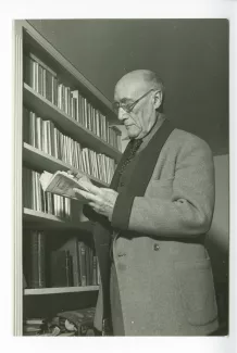 portrait photographique d’André Gide, avec lunettes, lisant un livre de Georges Simenon, debout à côté d’une bibliothèque, chez Richard Heyd, hiver 1947