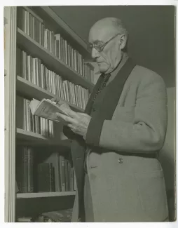 portrait photographique d’André Gide, avec lunettes, lisant un livre de Georges Simenon, debout à côté d’une bibliothèque, chez Richard Heyd, hiver 1947